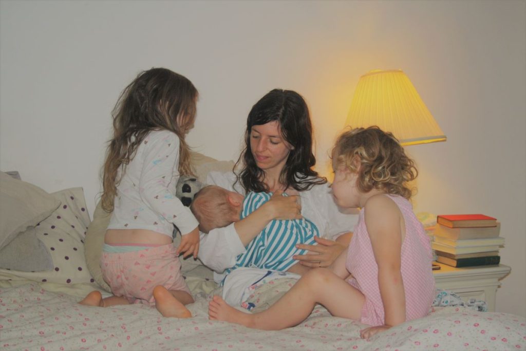 mother breastfeeding a baby in bed with two big sisters watching